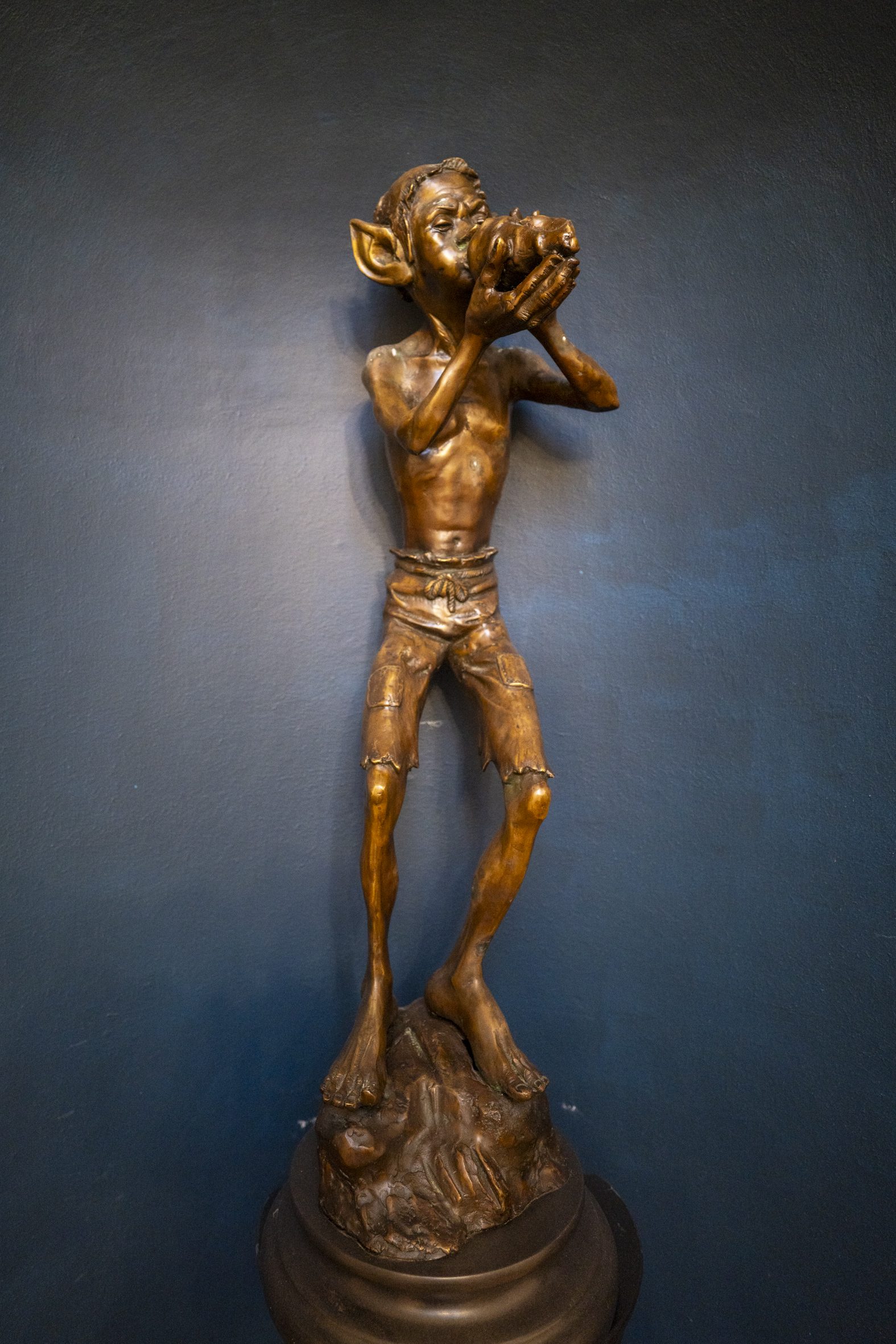 A bronze statue of a young boy with pointed ears, standing on a pedestal, holding a small fish-like object to his mouth.