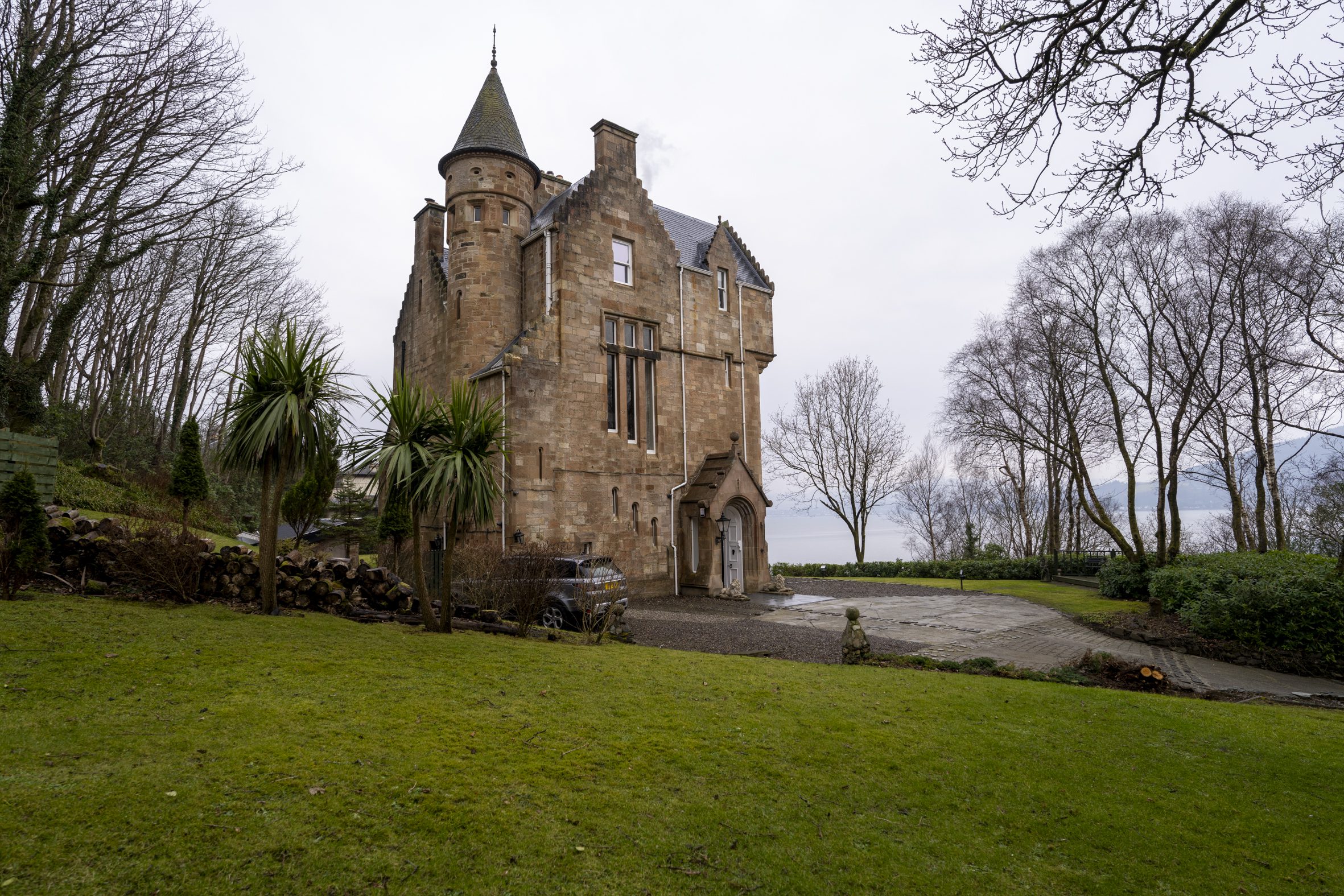 A historic Scottish castle, Cove Castle, nestled in lush greenery with palm-like plants in the foreground and a scenic view of Loch Long in the background.