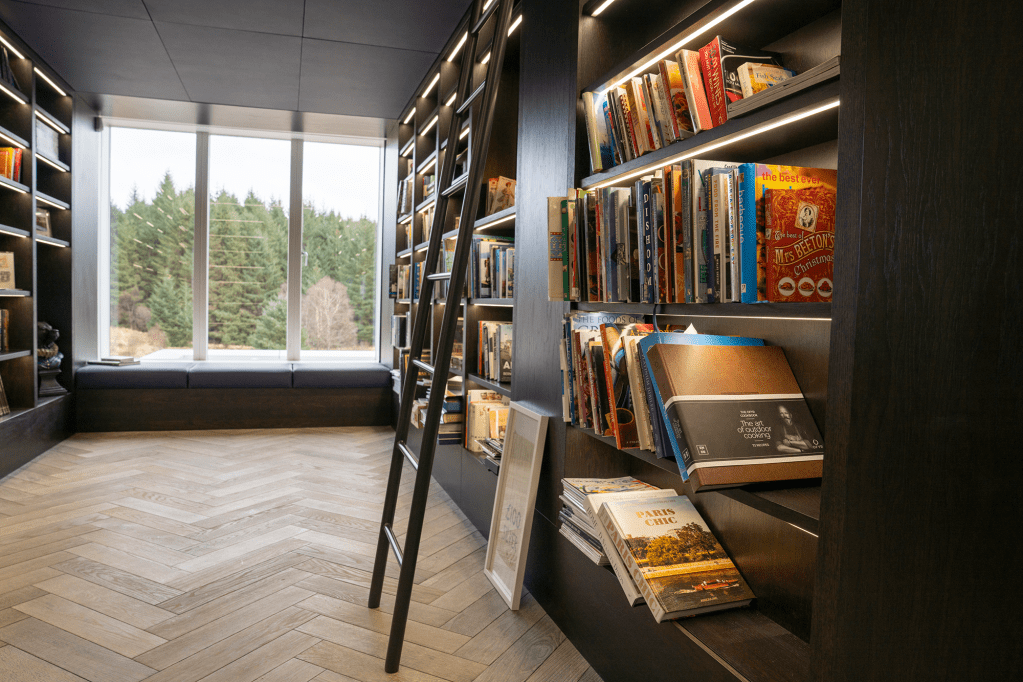 A cozy library interior with dark wooden shelves filled with books, a ladder for reaching higher shelves, and large windows overlooking a natural landscape.