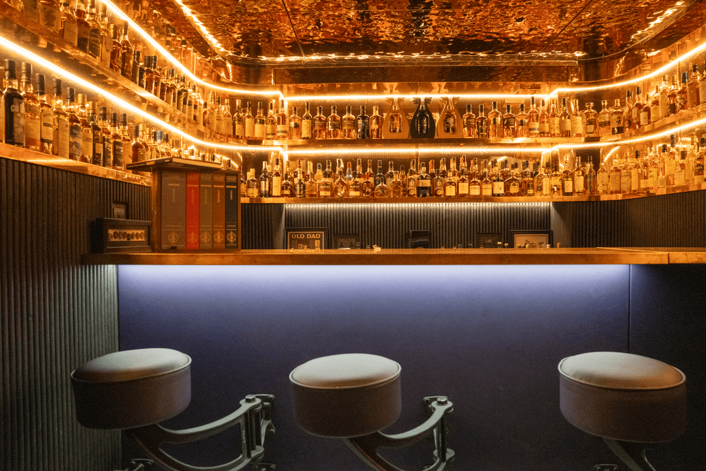 A stylish bar interior featuring a backlit shelf filled with various bottles of liquor, with modern bar stools in the foreground and warm lighting creating an inviting atmosphere.