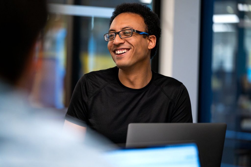A smiling man wearing glasses and a black shirt is seated at a table with laptops, appearing to engage in conversation in a modern office environment.