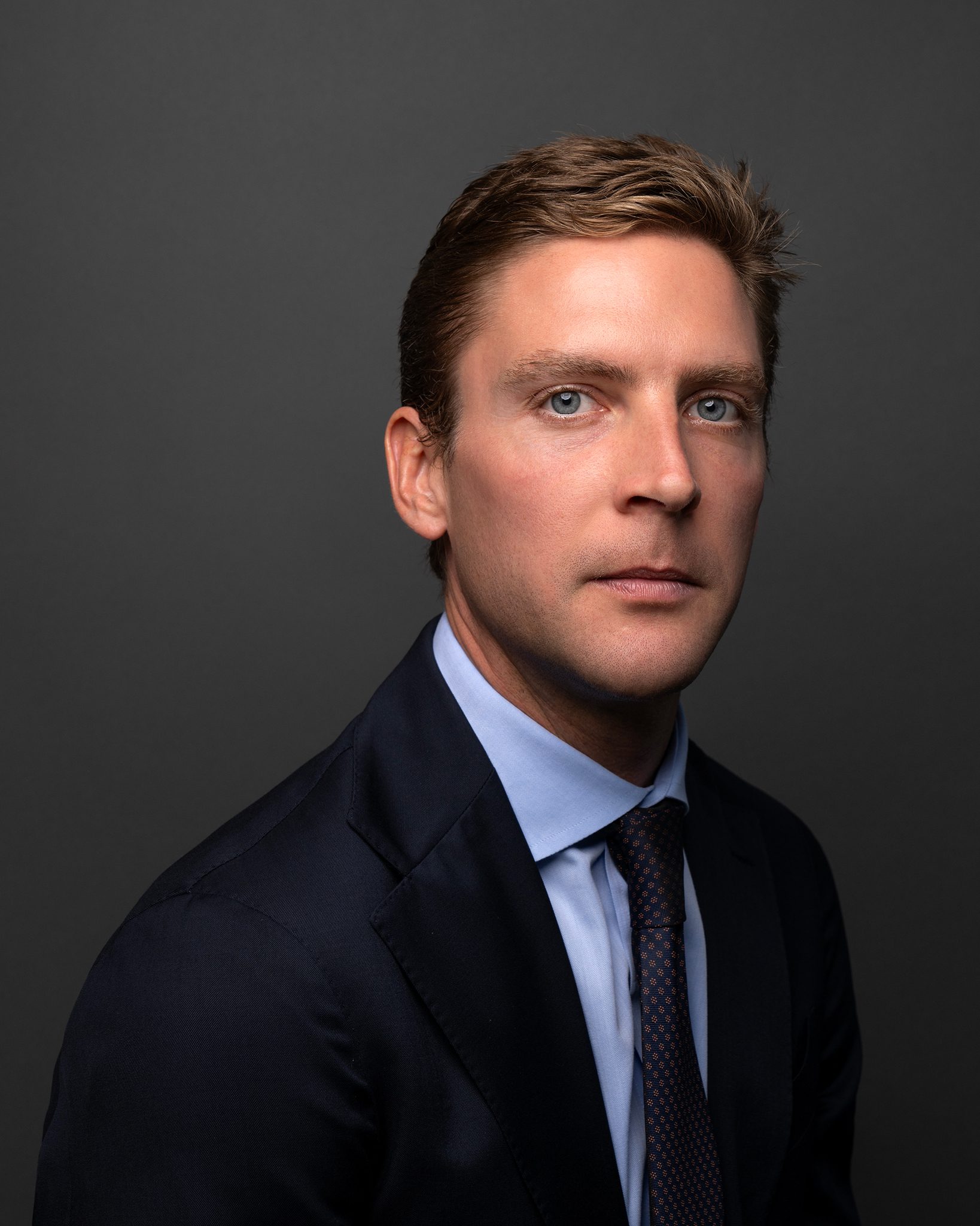 A professional portrait of a young man wearing a dark suit and a light blue shirt, seated against a dark gray background, looking directly at the camera with a serious expression.