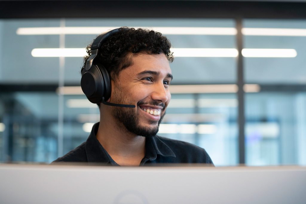 A smiling young man wearing a headset, sitting at a desk with a computer. The background features a modern office environment.