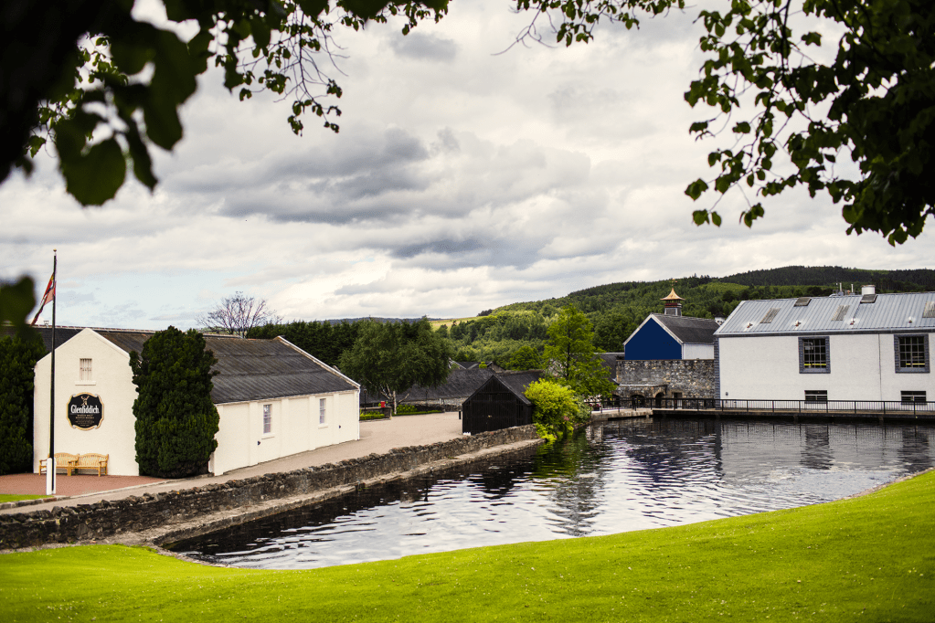 Wonderful Whisky 5 A scenic view of a Scottish whisky distillery situated by a peaceful body of water, surrounded by lush greenery and cloudy skies.