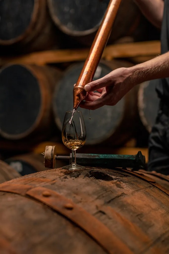 Wonderful Whisky 4 A person using a copper pipe to pour whisky into a glass placed on top of an old wooden barrel, with barrels in the background.