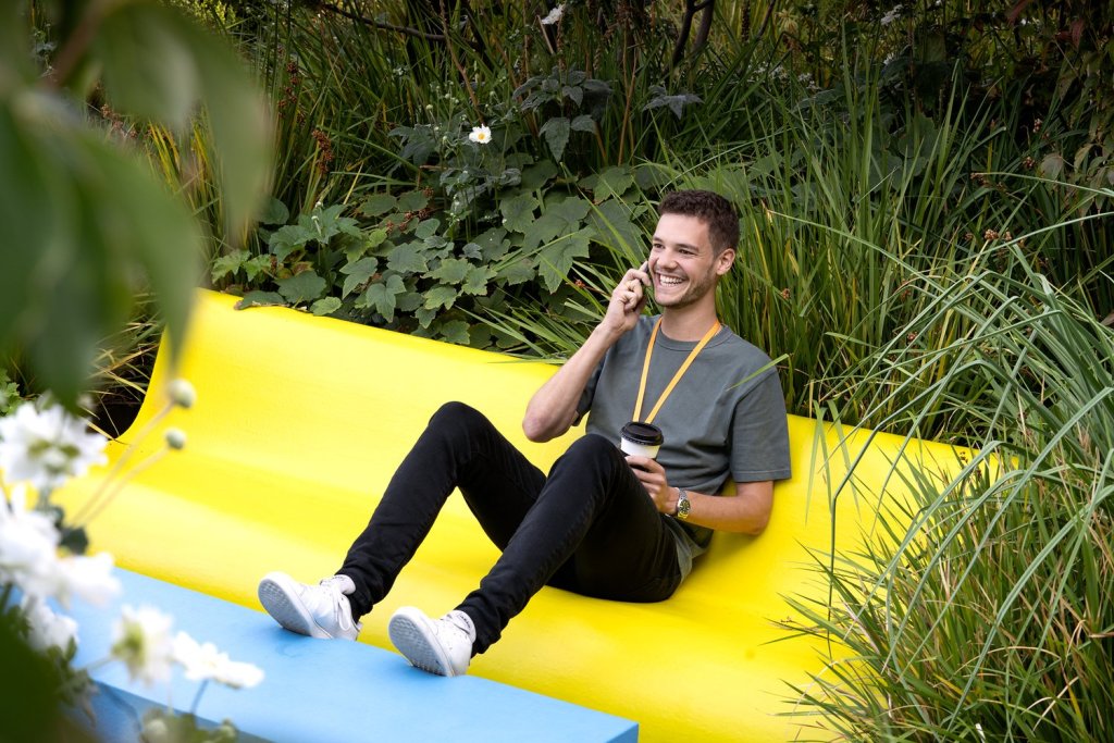 A smiling young man sitting on a yellow bench surrounded by greenery, holding a coffee cup and talking on the phone.
