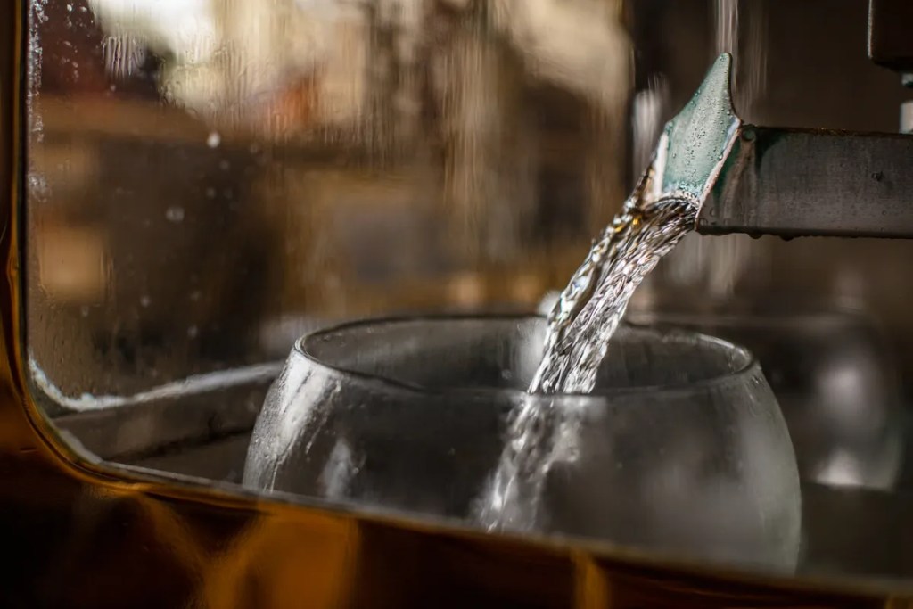 Wonderful Whisky 11 A close-up view of water being poured from a spout into a round glass vessel, with condensation visible on the glass.